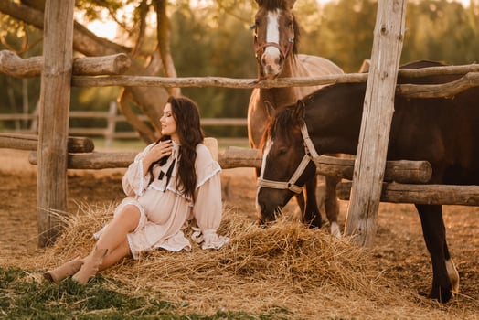 a pregnant woman in a dress in the countryside is sitting on the hay near the horses photo