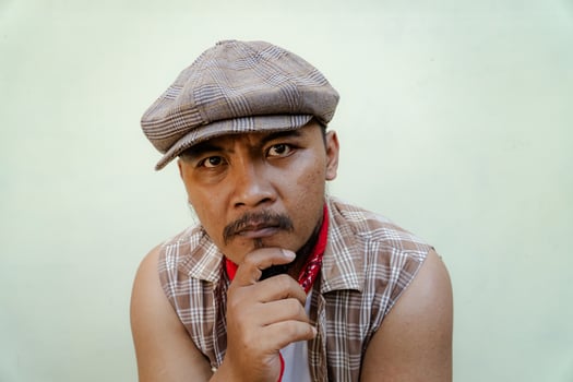 Half body portrait of adult Southeast Asian man in sleeveless shirt, bandana and newsboy hat. Macho male model with beard and mustache looking serious and fierce isolated on beige background. photo