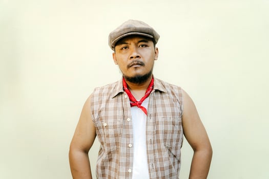 Half body portrait of adult Southeast Asian man in sleeveless shirt, bandana and newsboy hat. Macho male model with beard and mustache looking serious and fierce isolated on beige background. photo