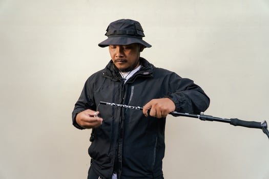 Photo of Asian man wearing adventuring outfit. Half body portrait of an adventurer with beard and mustache posing climber wearing jacket, boonie hat and trekking poles isolated on beige background