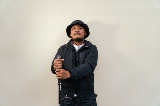 Photo of Asian man wearing adventuring outfit. Half body portrait of an adventurer with beard and mustache posing climber wearing jacket, boonie hat and trekking poles isolated on beige background