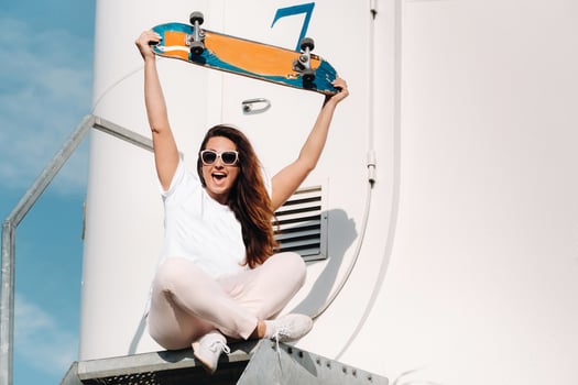 A girl in white clothes and glasses with a skate in her hands is photographed near large wind turbines in a field with trees.Modern woman with a riding Board in a field with windmills photo