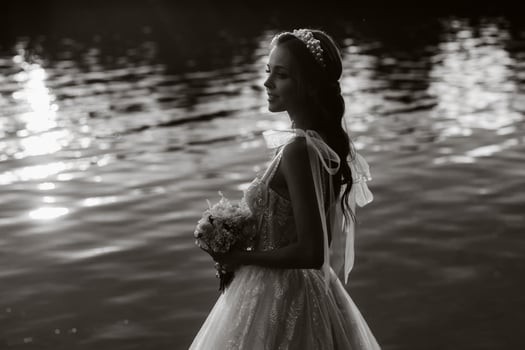 An elegant bride in a white dress and gloves stands by the river in the Park with a bouquet, enjoying nature at sunset.A model in a wedding dress and gloves in a natur. black and white photo. photo