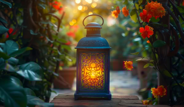 A lantern with a candle inside is lit up in a garden. The lantern is blue and has a gold design. The garden is filled with orange flowers and green plants. The lantern is placed on a brick ledge photo