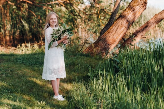 A beautiful nine-year-old blonde girl with long hair in a long white dress, holding a bouquet of lilies of the valley flowers, walking in nature in the Park.Summer, sunset. photo