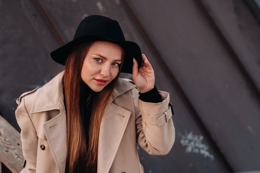 A stylish young woman in a beige coat and black hat sits on a rooftop in the city center. Women's street fashion. Autumn clothing.Urban style photo