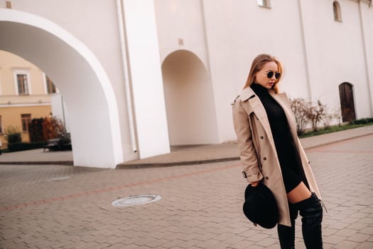 Stylish young woman in a beige coat and a black hat in his hands and glasses on a city street. Women's street fashion. Autumn clothing.Urban style. photo