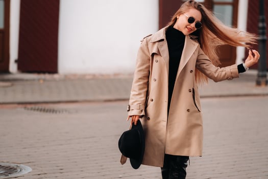 Stylish young woman in a beige coat and a black hat in his hands and glasses on a city street. Women's street fashion. Autumn clothing.Urban style. photo