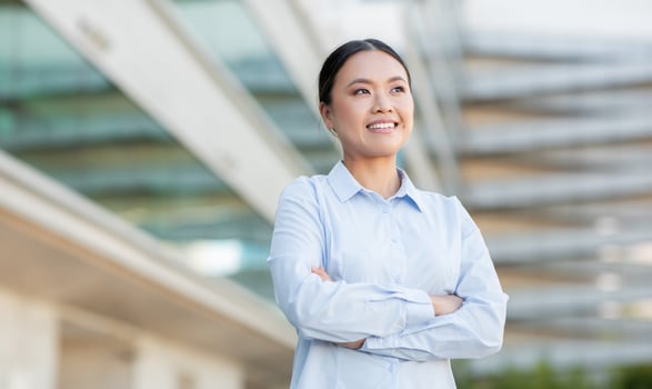 A confident Asian woman stands outdoors with her arms crossed, smiling and enjoying the sunshine. The backdrop features a sleek modern building, hinting at a professional environment. photo