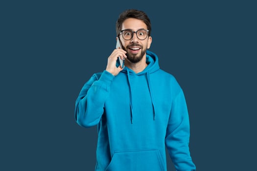 A young man in a bright blue hoodie excitedly talks on the phone, with a wide smile and enthusiastic demeanor, standing in front of a solid blue background. photo