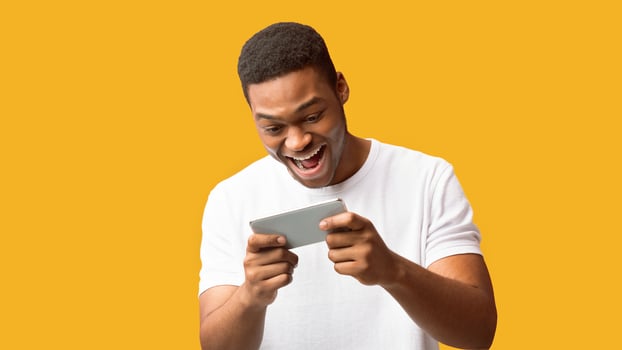 Online Game App. Happy black guy playing games on smartphone over orage background, copyspace, panorama photo