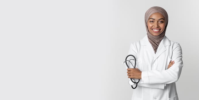 Portrait Of Young Afro Muslim Female Doctor In Hijab Wearing White Coat And Holding Stethoscope, Posing With Folded Arms Over Light Background photo