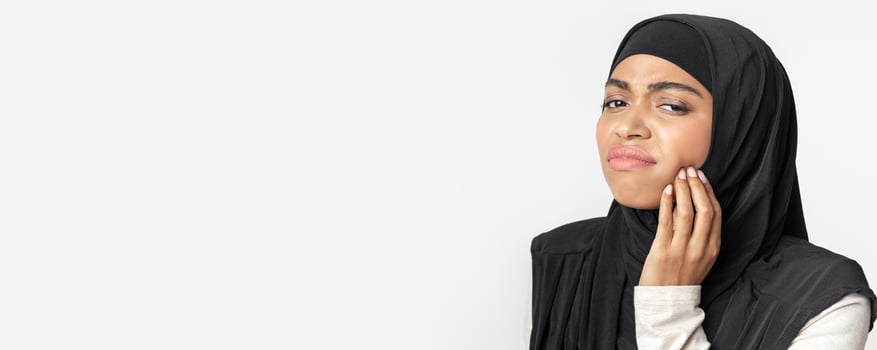 Toothache concept. Black muslim woman in headscarf suffering from teeth pain, touching her inflamed cheek, gray background with free space, panorama photo