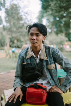 A Southeast Asian man wearing a denim jacket, sitting on a bench in a city square, photo