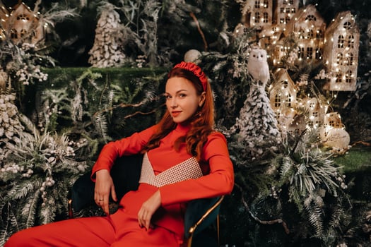 a girl in a red suit is sitting on a chair near the Christmas background. a woman in red on the background of Christmas trees and small Christmas houses photo