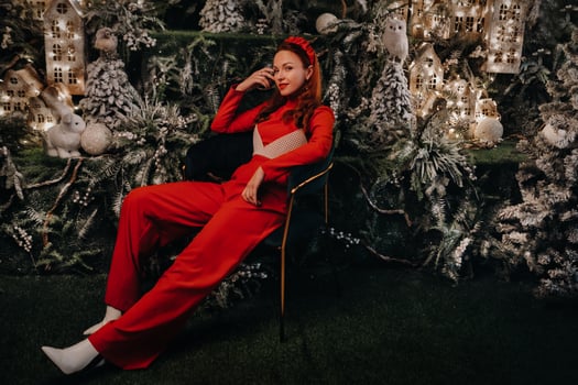 a girl in a red suit is sitting on a chair near the Christmas background. a woman in red on the background of Christmas trees and small Christmas houses photo