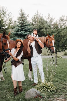 A family in white clothes with their son stand near two beautiful horses in nature. A stylish couple with a child are photographed with horses photo