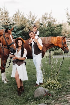 A family in white clothes with their son stand near two beautiful horses in nature. A stylish couple with a child are photographed with horses photo