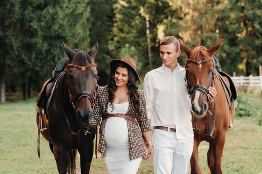 a pregnant girl in a hat and her husband in white clothes stand next to horses in the forest in nature.Stylish pregnant woman with a man with horses.Family. photo