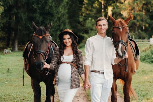 a pregnant girl in a hat and her husband in white clothes stand next to horses in the forest in nature.Stylish pregnant woman with a man with horses.Family. photo