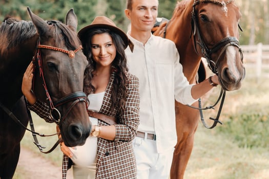 a pregnant girl in a hat and her husband in white clothes stand next to horses in the forest in nature.Stylish pregnant woman with a man with horses.Family. photo
