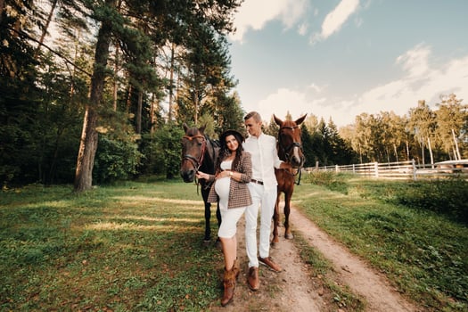 a pregnant girl in a hat and her husband in white clothes stand next to horses in the forest in nature.Stylish pregnant woman with a man with horses.Family. photo