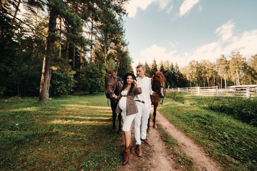 a pregnant girl in a hat and her husband in white clothes stand next to horses in the forest in nature.Stylish pregnant woman with a man with horses.Family. photo