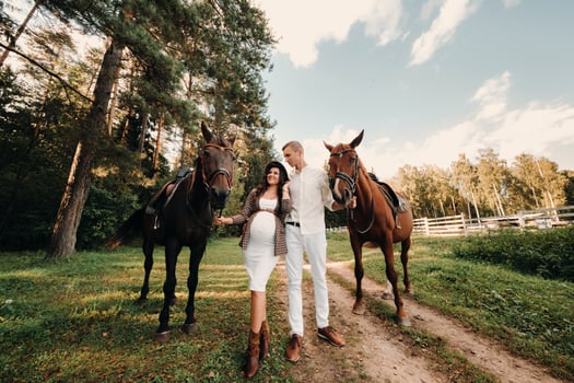 a pregnant girl in a hat and her husband in white clothes stand next to horses in the forest in nature.Stylish pregnant woman with a man with horses.Family. photo