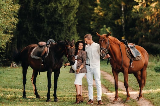 a pregnant girl in a hat and her husband in white clothes stand next to horses in the forest in nature.Stylish pregnant woman with a man with horses.Family. photo