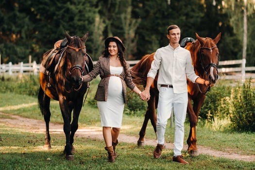 a pregnant girl in a hat and her husband in white clothes stand next to horses in the forest in nature.Stylish pregnant woman with a man with horses.Family. photo