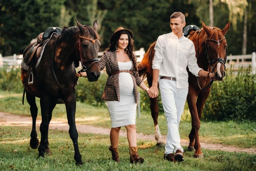 A pregnant woman in a hat with a man in white clothes walking with horses in nature. A family waiting for a child walks in the woods. photo