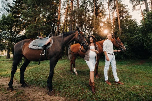 a pregnant girl in a hat and her husband in white clothes stand next to horses in the forest in nature.Stylish pregnant woman with a man with horses.Family. photo
