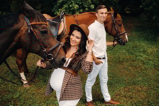 A pregnant woman in a hat with a man in white clothes walking with horses in nature. A family waiting for a child walks in the woods photo