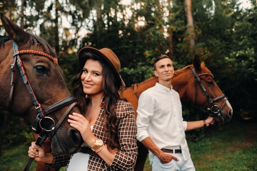 a pregnant girl in a hat and a man in white clothes stand next to horses in the forest in nature.Stylish pregnant woman with her husband with horses.Married couple. photo