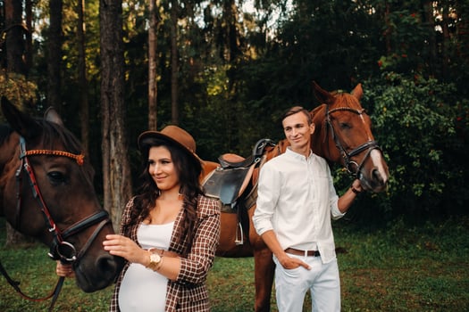 a pregnant girl in a hat and a man in white clothes stand next to horses in the forest in nature.Stylish pregnant woman with her husband with horses.Married couple. photo