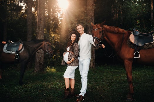 a pregnant girl in a hat and her husband in white clothes stand next to horses in the forest in nature.Stylish pregnant woman with a man with horses.Family. photo