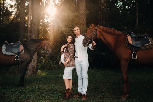 a pregnant girl in a hat and her husband in white clothes stand next to horses in the forest in nature.Stylish pregnant woman with a man with horses.Family. photo