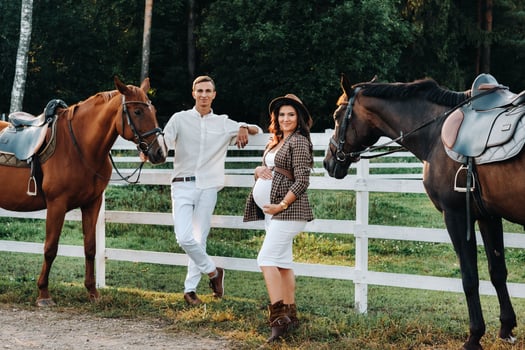 a pregnant girl in a hat and a man in white clothes stand next to horses near a white fence.Stylish pregnant woman with a man with horses.Married couple. photo