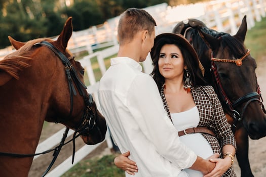 a pregnant girl in a hat and her husband in white clothes stand next to the horses near the horse corral.Stylish pregnant woman with a man with horses.Family photo