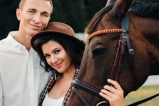 a pregnant girl in a hat and a man in white clothes stand next to horses at a White fence.Stylish family waiting for a child strolling in nature photo