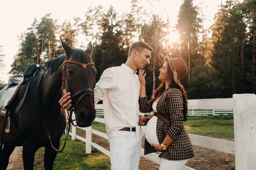 a pregnant girl in a hat and a man in white clothes stand next to horses at a White fence.Stylish family waiting for a child strolling in nature photo