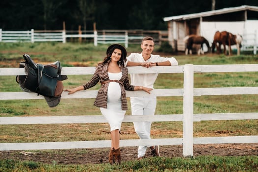a pregnant girl in a hat and her husband in white clothes stand next to the horse corral.a stylish couple waiting for a child stand on the street near the horse corral. photo