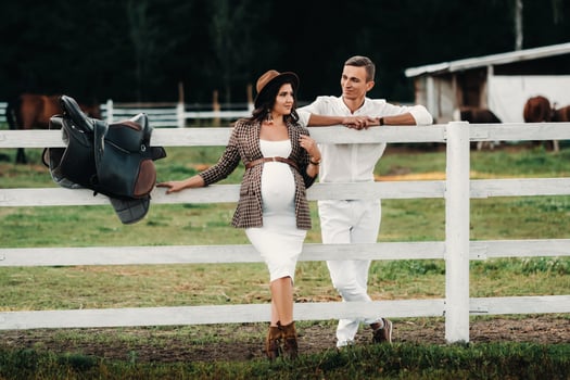 a pregnant girl in a hat and her husband in white clothes stand next to a horse corral at sunset.a stylish couple is waiting for a child in nature photo