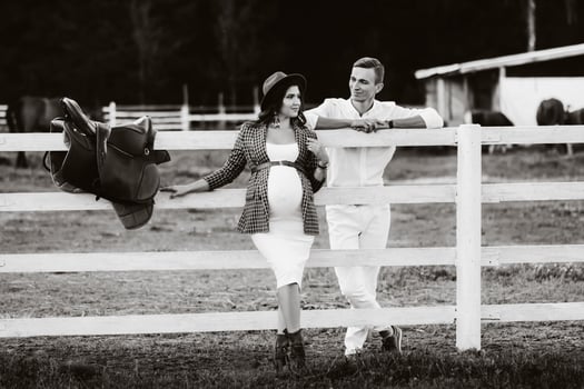 a pregnant girl in a hat and her husband in white clothes stand next to the horse corral.a stylish couple waiting for a child stand on the street near the horse corral.black and white photo