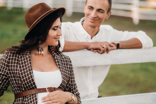 a pregnant girl in a hat and her husband in white clothes stand next to a horse corral at sunset.a stylish couple is waiting for a child in nature photo
