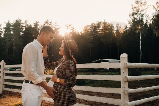 a pregnant girl in a hat and her husband in white clothes stand next to a horse corral at sunset.a stylish couple is waiting for a child in nature photo