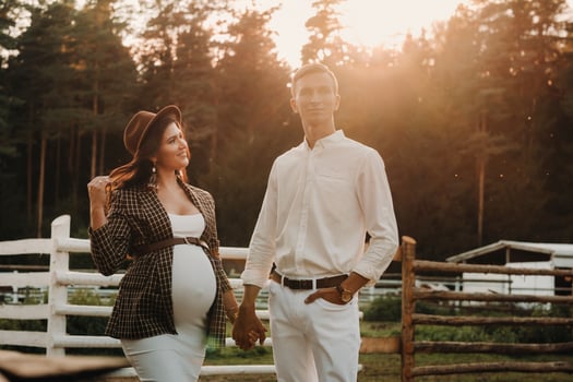 a pregnant girl in a hat and her husband in white clothes stand next to a horse corral at sunset.a stylish couple is waiting for a child in nature photo
