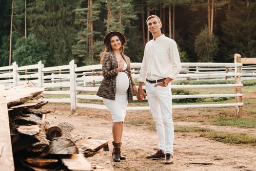 a pregnant girl in a hat and her husband in white clothes stand next to a horse corral at sunset.a stylish couple is waiting for a child in nature photo
