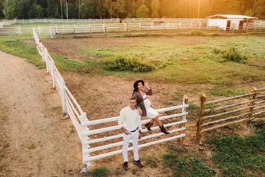 a pregnant girl in a hat and her husband in white clothes stand next to a horse corral at sunset.a stylish couple is waiting for a child in nature photo