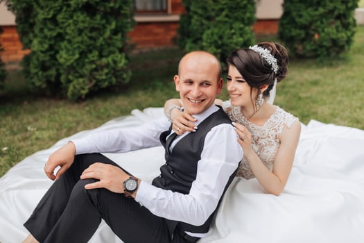 A man and woman are sitting on a blanket in a grassy area. The man is wearing a watch and the woman is wearing a wedding dress. They are smiling and seem to be enjoying each other's company photo